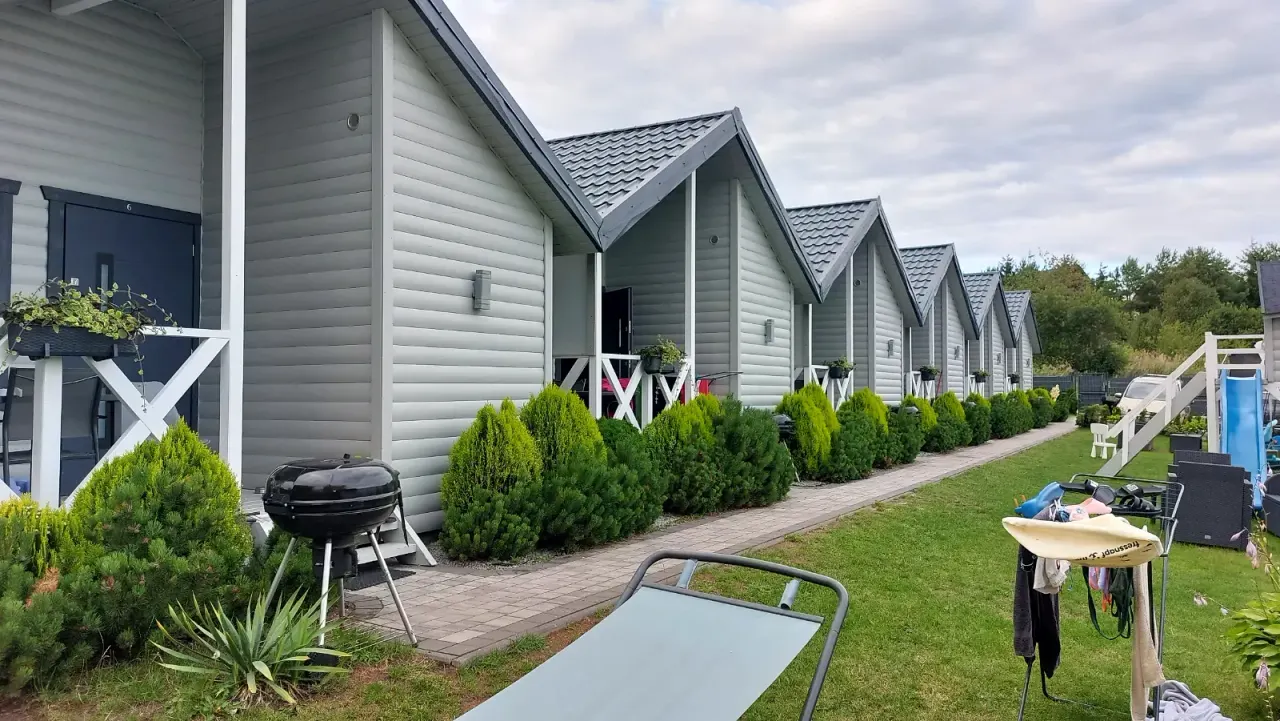 BookNoFees : location de vacances à Jarosławiec, Pologne – Une rangée de chalets en bois gris dotés de porches, d'un barbecue et d'une pelouse verte