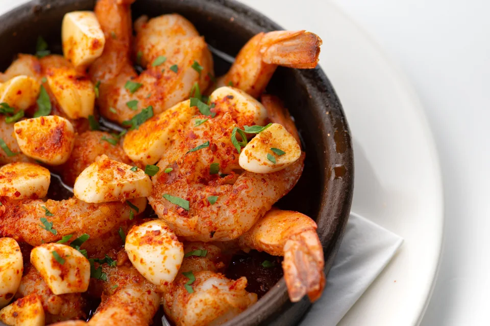 BookNoFees: Photo d’illustration : Crevettes à l'ail garnies d'herbes hachées, servies dans un bol noir posé sur une assiette blanche